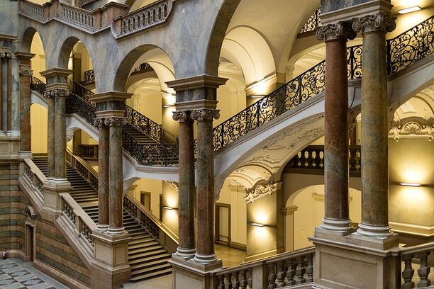L’immagine mostra l’interno di un grande edificio storico con colonne, archi e una scalinata monumentale. L’architettura elegante e articolata suggerisce un luogo istituzionale o culturale. Nel contesto dell’articolo, l’immagine richiama simbolicamente i processi attraverso cui le istituzioni, la cultura e il potere contribuiscono alla costruzione delle narrazioni storiche e della memoria collettiva.