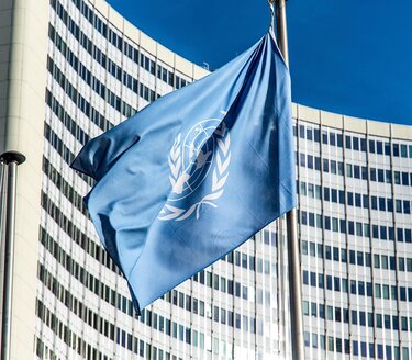 L’immagine mostra la bandiera delle Nazioni Unite che sventola su un’asta davanti a un grande edificio moderno con numerose finestre. Il simbolo dell’ONU è visibile al centro della bandiera. L’immagine richiama il contesto delle istituzioni internazionali e il tema del diritto internazionale, al centro della riflessione sull’equilibrio tra sovranità degli stati e misure di sanzione unilaterale.