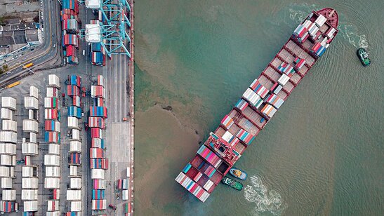 L’immagine mostra una nave portacontainer vista dall’alto mentre naviga accanto a un grande porto commerciale pieno di container disposti in file ordinate. Le gru portuali e le infrastrutture logistiche indicano un importante nodo dei traffici marittimi internazionali. Nel contesto dell’articolo, l’immagine richiama il tema delle restrizioni economiche e commerciali legate alle misure di embargo e alle loro possibili conseguenze economiche e umanitarie.