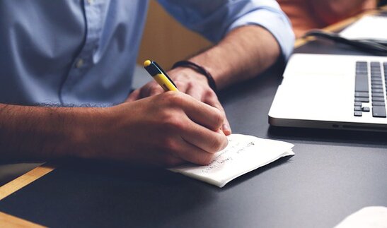 L’immagine mostra una persona seduta a una scrivania mentre scrive appunti su un piccolo quaderno con una matita. Accanto al quaderno si trova un computer portatile aperto. La scena richiama un momento di studio, analisi e riflessione. Nel contesto dell’articolo, l’immagine rappresenta il lavoro di ricerca e di elaborazione metodologica che sta alla base del progetto “Internazionale per la Pace”.