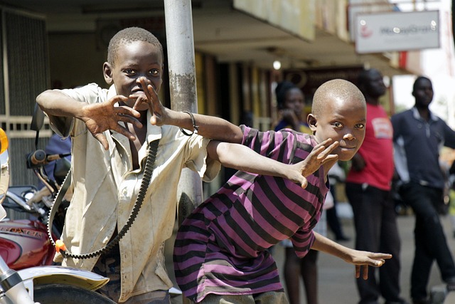 L’immagine mostra due ragazzi africani in primo piano che guardano verso la macchina fotografica con gesti dinamici delle mani, mentre sullo sfondo si intravede un contesto urbano con altre persone. L’atmosfera è vivace e spontanea. Nel progetto espositivo questa immagine rappresenta simbolicamente il popolo ospite che, in ogni edizione dell’iniziativa, porta la propria voce, la propria cultura e la propria esperienza storica all’interno del percorso.